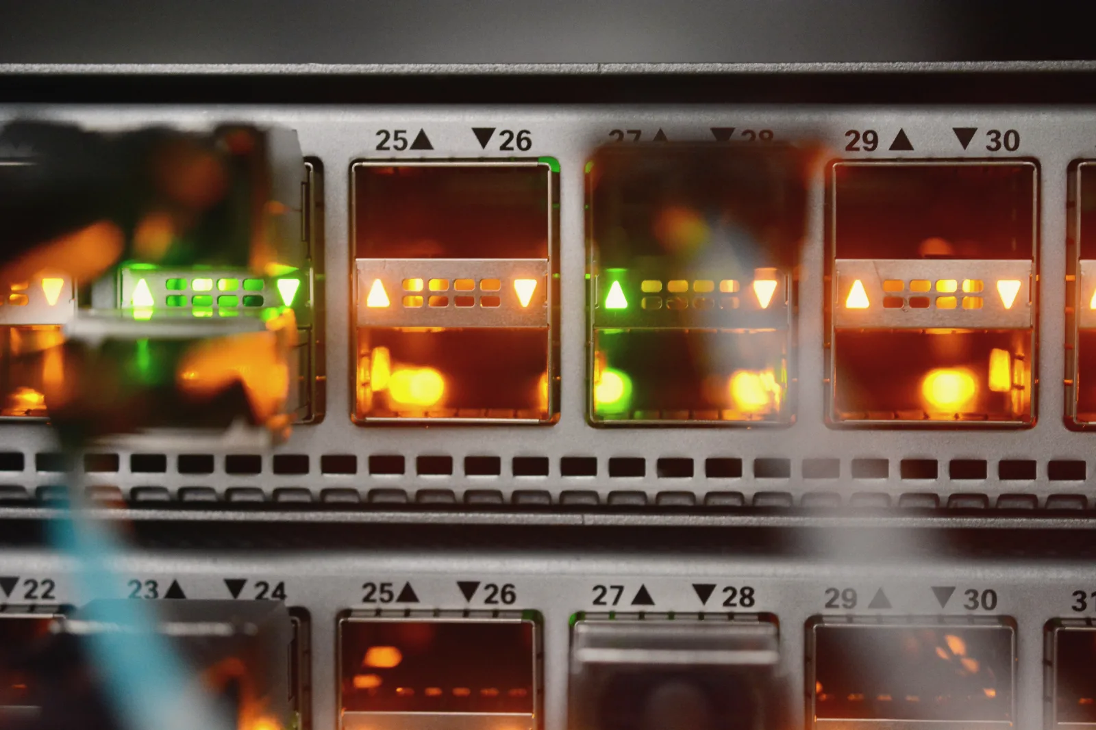 Close-up of a network switch: a row of square ports glows orange and green under printed port numbers, with blurred plugs and cables in the foreground.