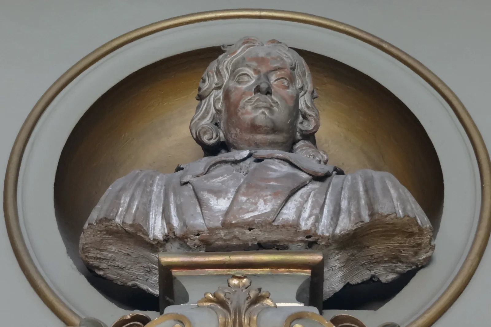 Seen from below, a bust of Pierre de Fermat with long curled hair and a wide collar, its gray paint worn away in reddish patches, stands in a round niche with a gold rim above a pedestal with gilded trim.