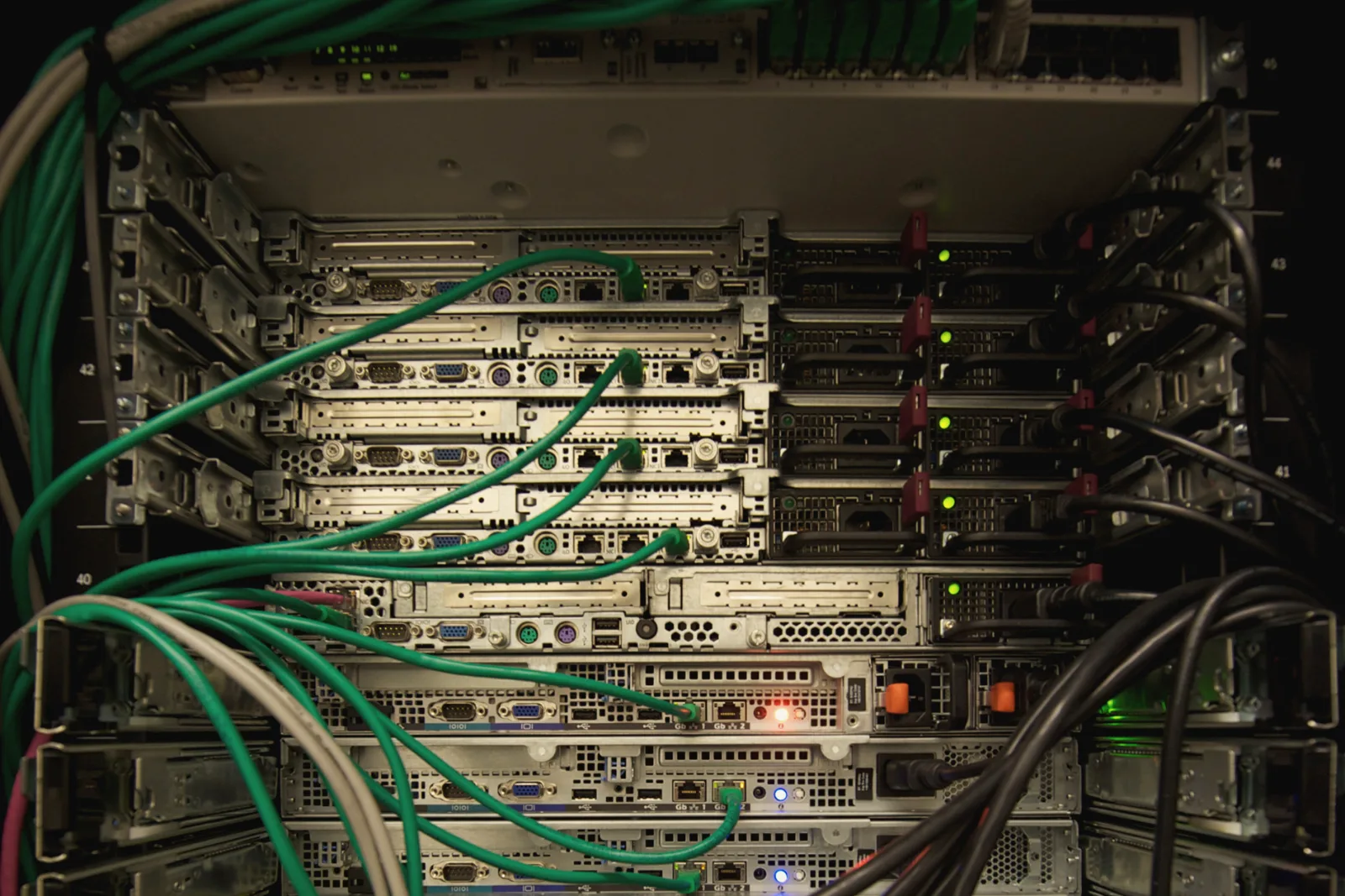 The back of a server rack in the dark: a stack of metal servers with small green, orange and blue lights, crossed by bundles of green, gray and black cables.