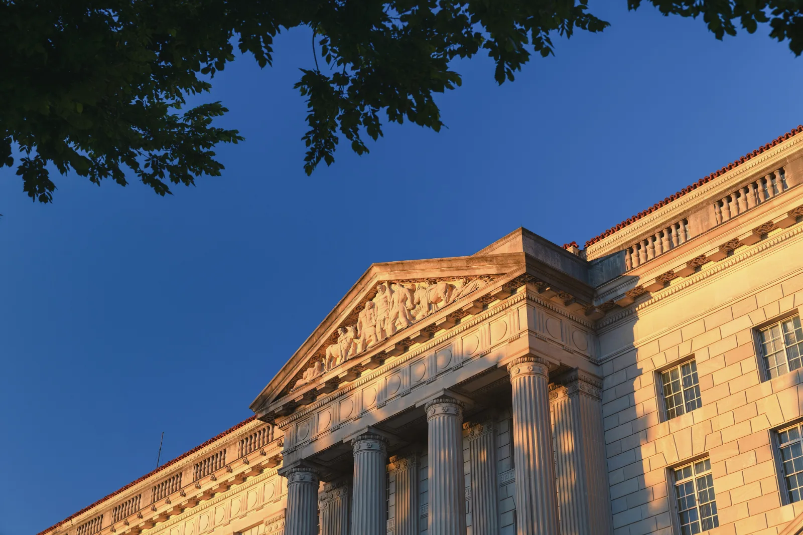 The stone pediment and columns of a large neoclassical government building glow orange in low sunlight against a deep blue sky, under dark tree branches.