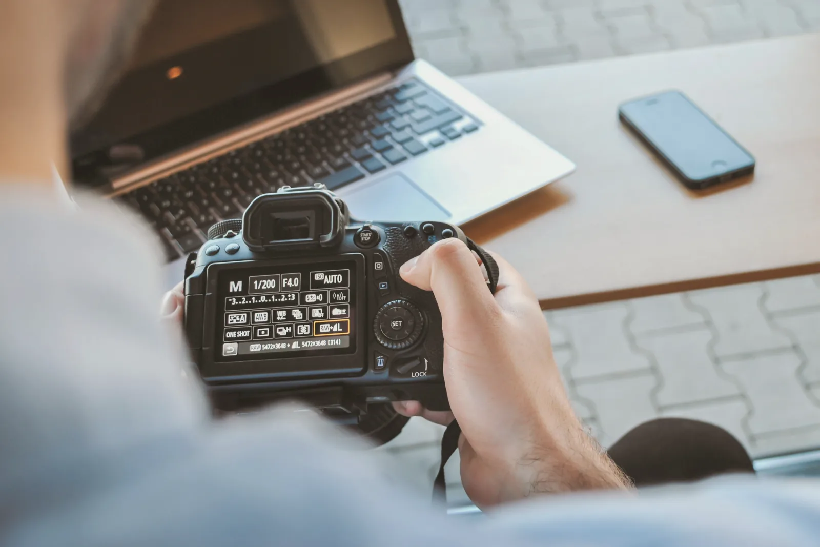 Seen over a person's shoulder, two hands hold a digital camera whose rear screen shows shooting settings, above a table with an open laptop and a smartphone.