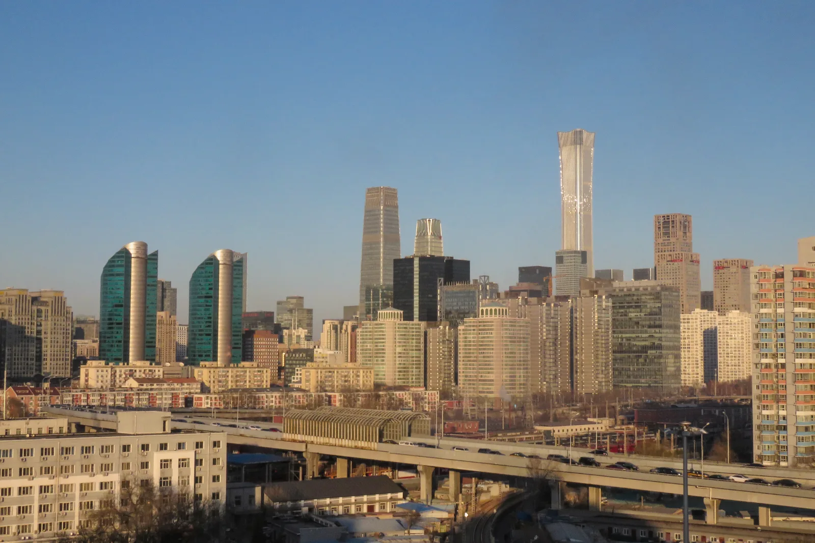 Glass and stone skyscrapers, one much taller than the rest, rise above low apartment blocks and an elevated road under a clear blue sky.