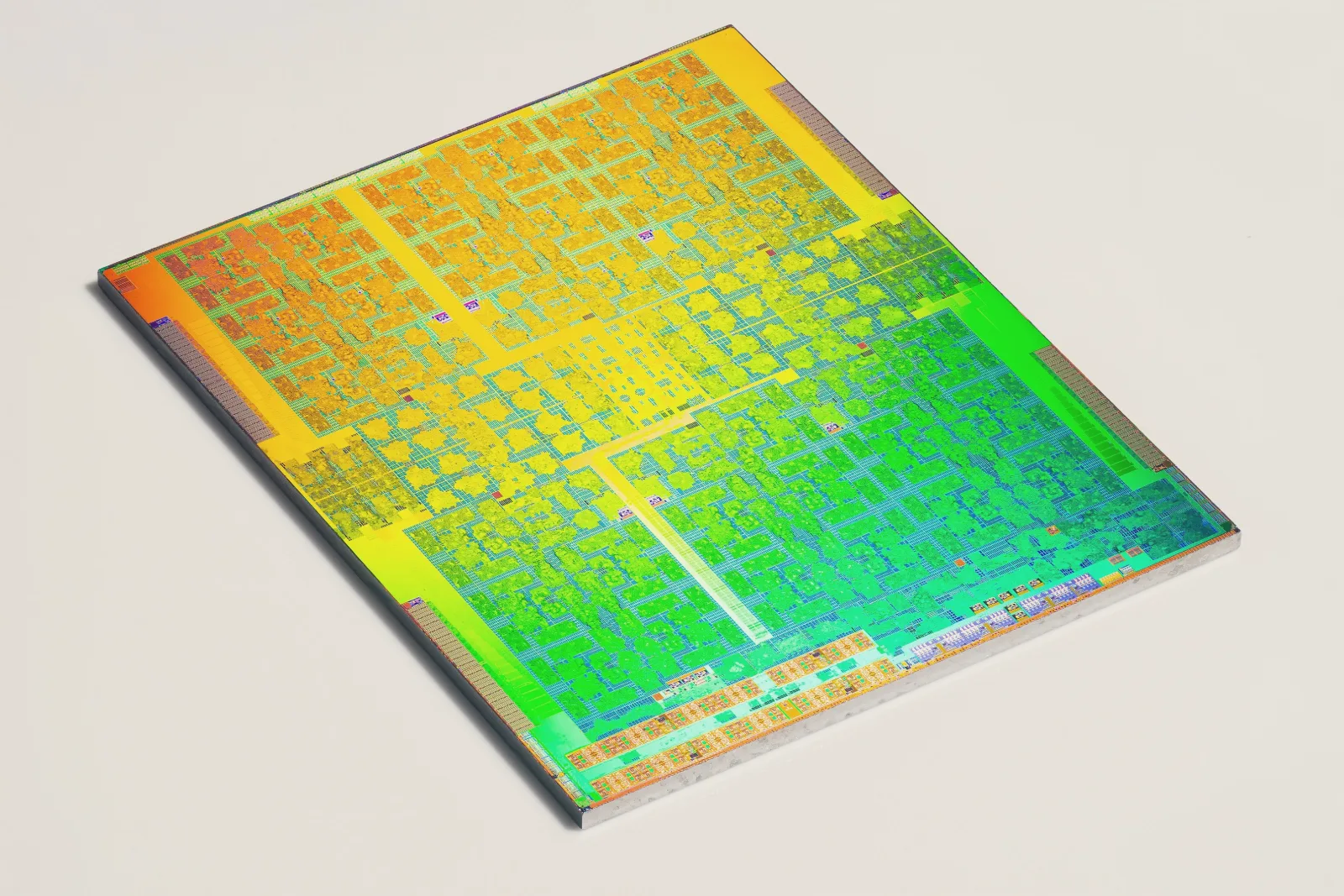 The bare silicon die of a large graphics processor lies at an angle on a white surface, its circuitry shimmering orange, yellow and green.
