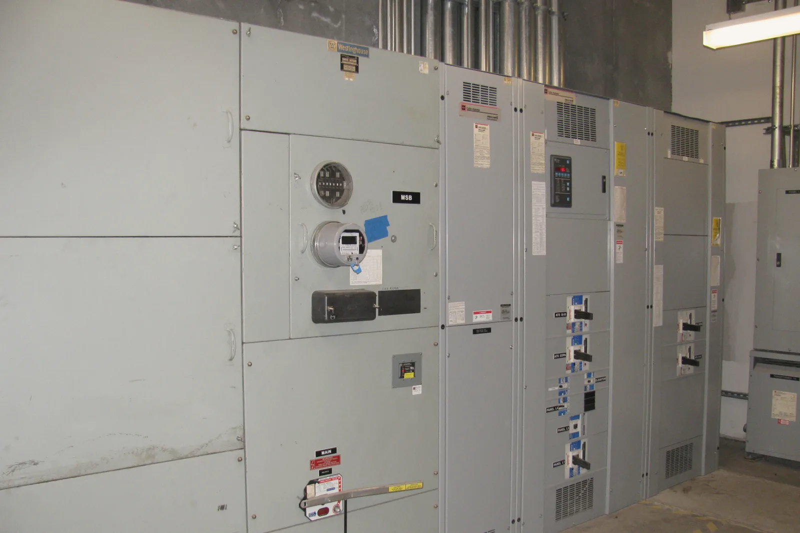 A row of gray switchgear cabinets stands in an electrical room, with metal conduits rising to the ceiling. One cabinet carries a round, glass-covered electricity meter with a digital display and a blue tag, below a second round fitting.