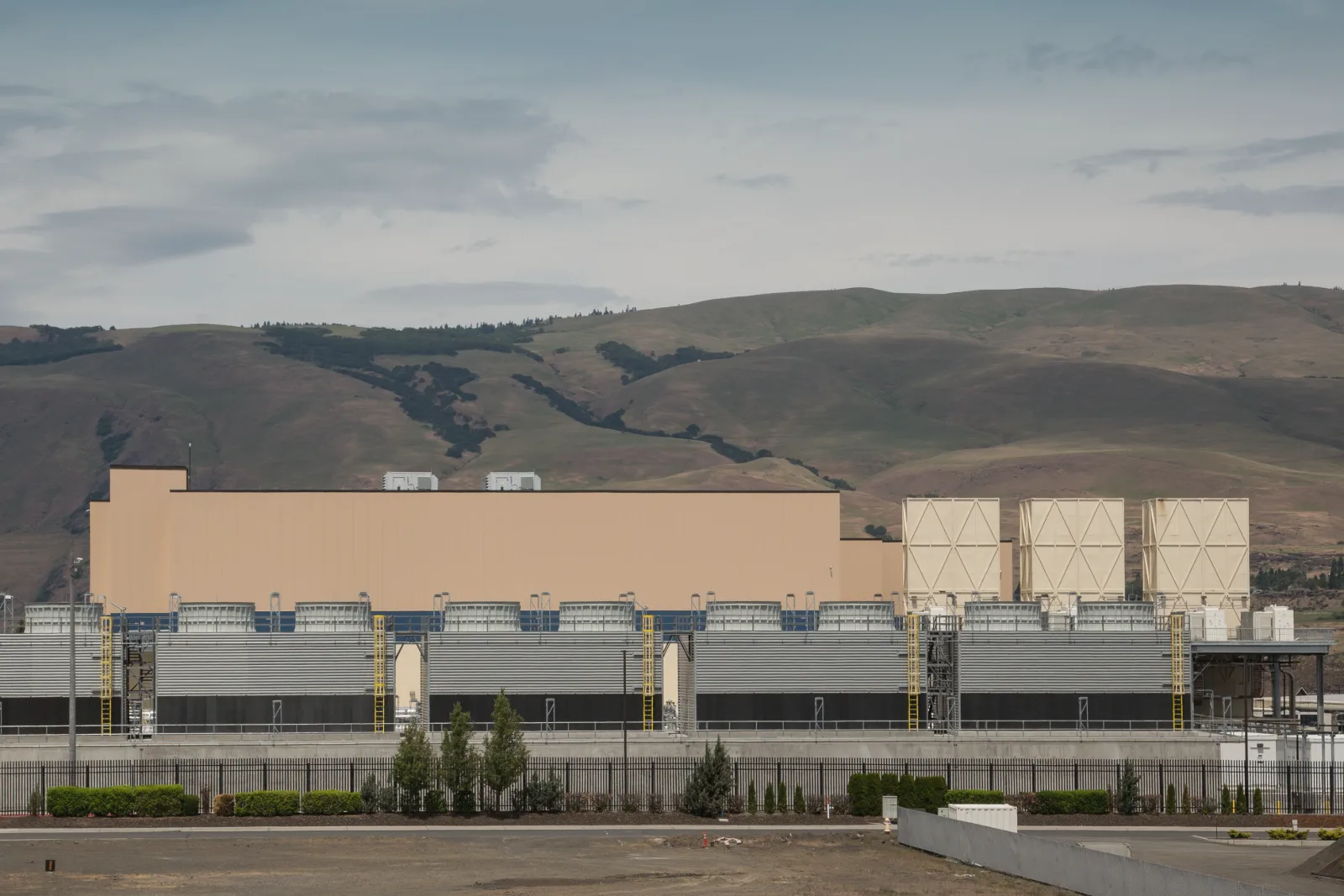 A long beige data center building stands behind a row of gray industrial units and a black fence, with brown and green hills under a cloudy sky behind it.