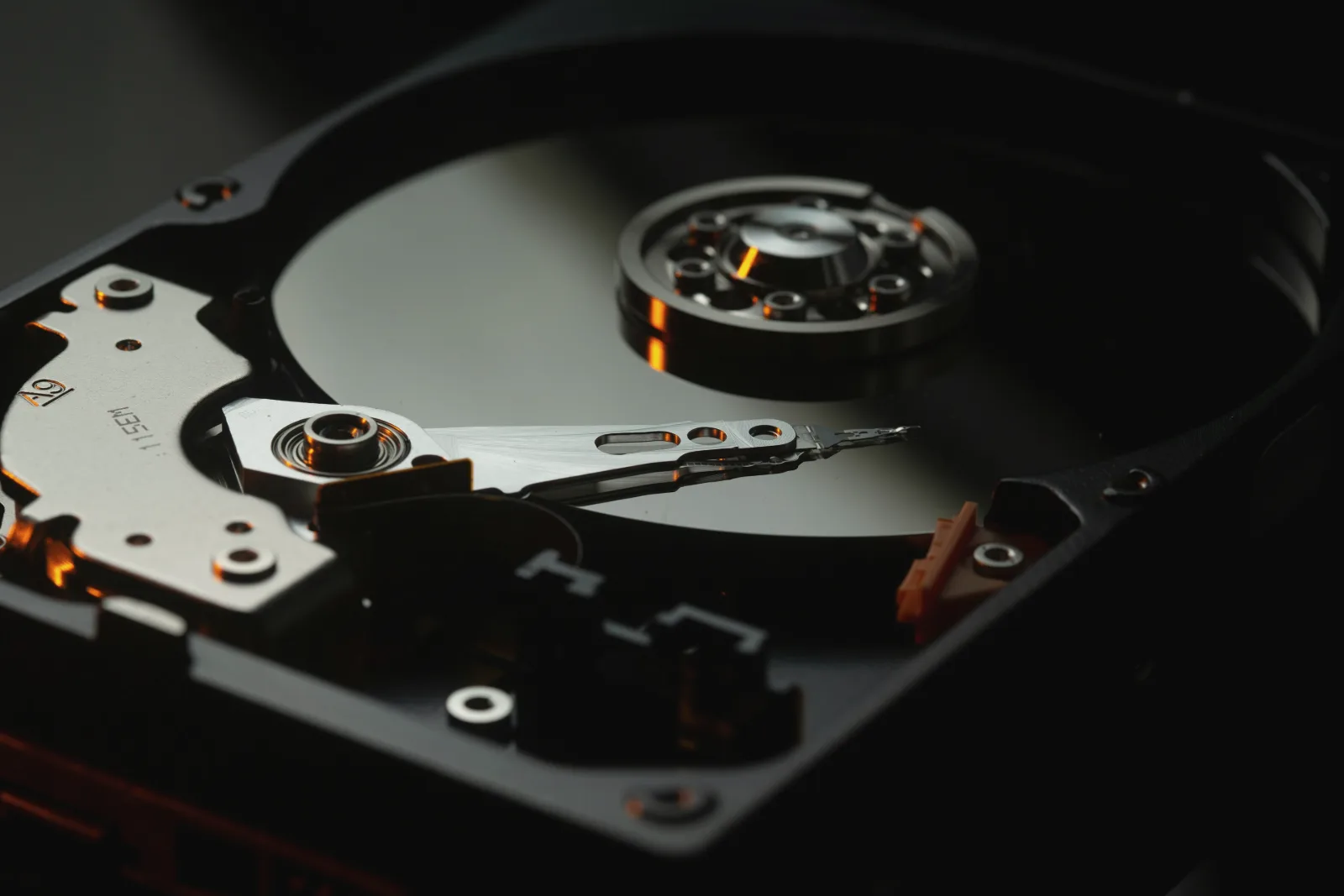 The inside of an opened hard disk drive in dim light, its mirror-like platter, spindle and read arm catching orange highlights.