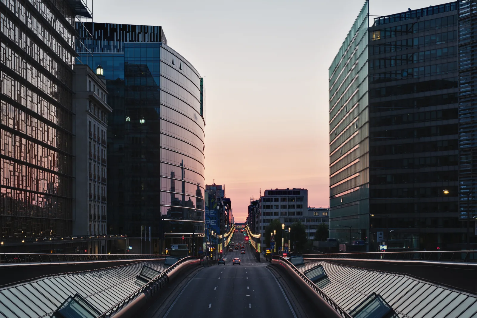 A wide road runs between tall glass office buildings toward a pink and orange evening sky, with streetlights and a few cars far down the street.