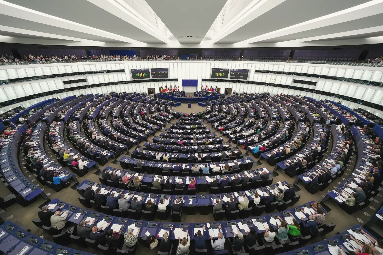 A wide view of the European Parliament's semicircular chamber, its rows of blue desks filled with members voting, under a white ceiling, with the EU flag and national flags behind the presiding officers.
