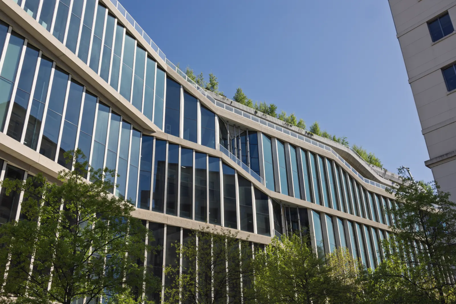 The glass and pale stone facade of a long office building curves in waves under a clear blue sky, with trees on its roof terraces and in the street below.