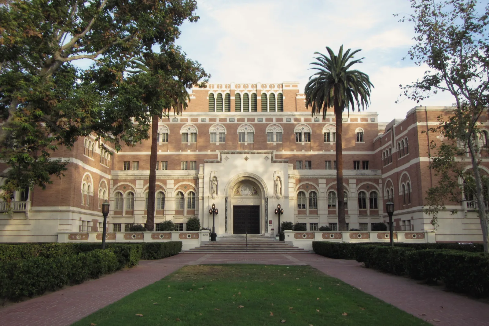 A large red-brick and white-stone library with an arched central entrance stands behind a tall palm tree, leafy trees, a lawn and brick paths in warm sunlight.