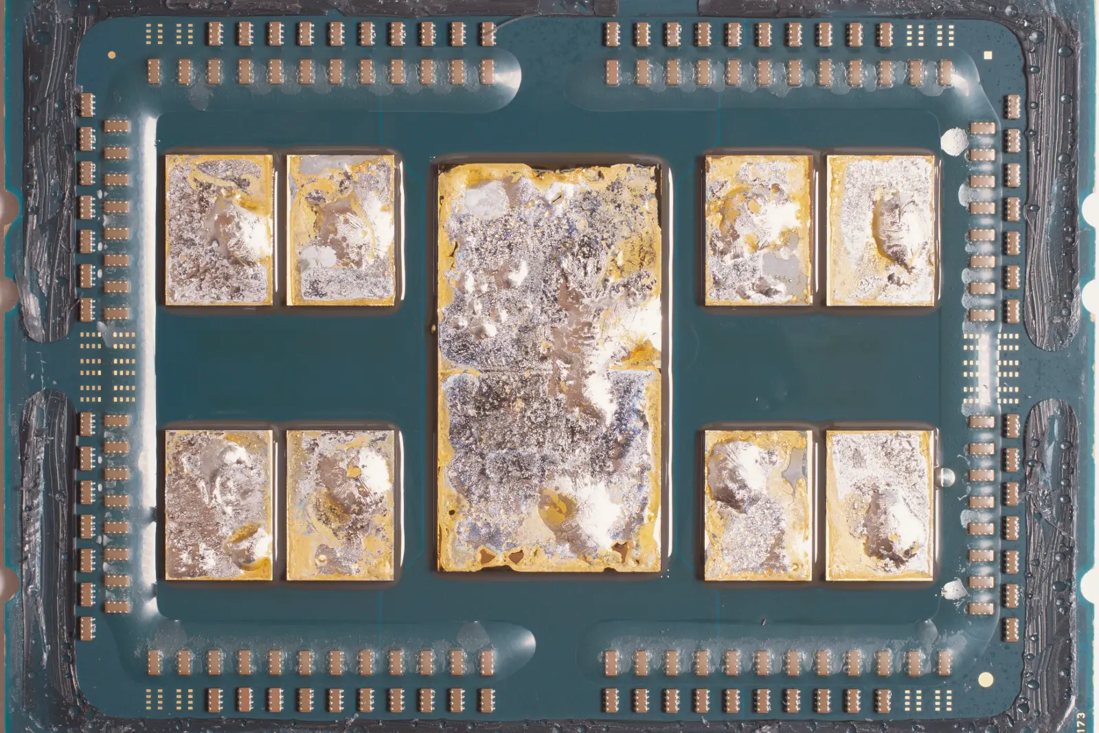 A server processor with its lid removed: eight small chips and one large central chip, covered in silvery solder residue, sit on a teal circuit board ringed by rows of tiny components.