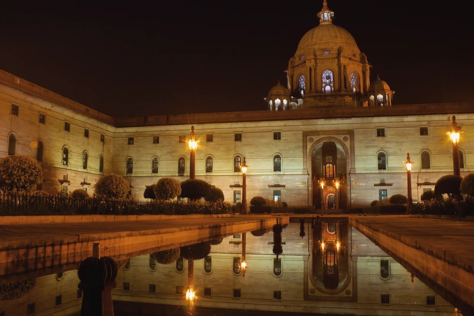 A long sandstone building with a central dome, lit by lamps at night, is reflected in a still pool in the foreground.