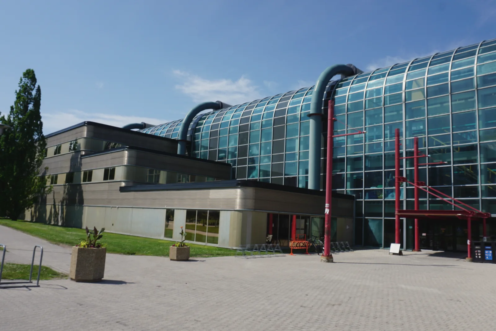滑铁卢大学的 Davis Centre 大楼:弧形的玻璃幕墙上方弯着几根粗大的灰色管道,入口处立着红色立柱;楼前是铺砖广场和混凝土花坛,天空湛蓝。