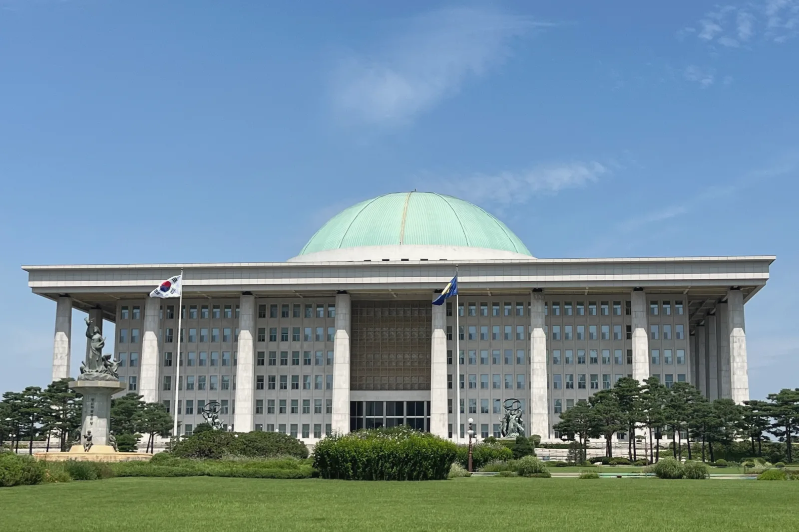 A wide white parliament building with a green dome and a row of tall square columns stands behind a lawn and pine trees, with two flags in front, under a blue sky.