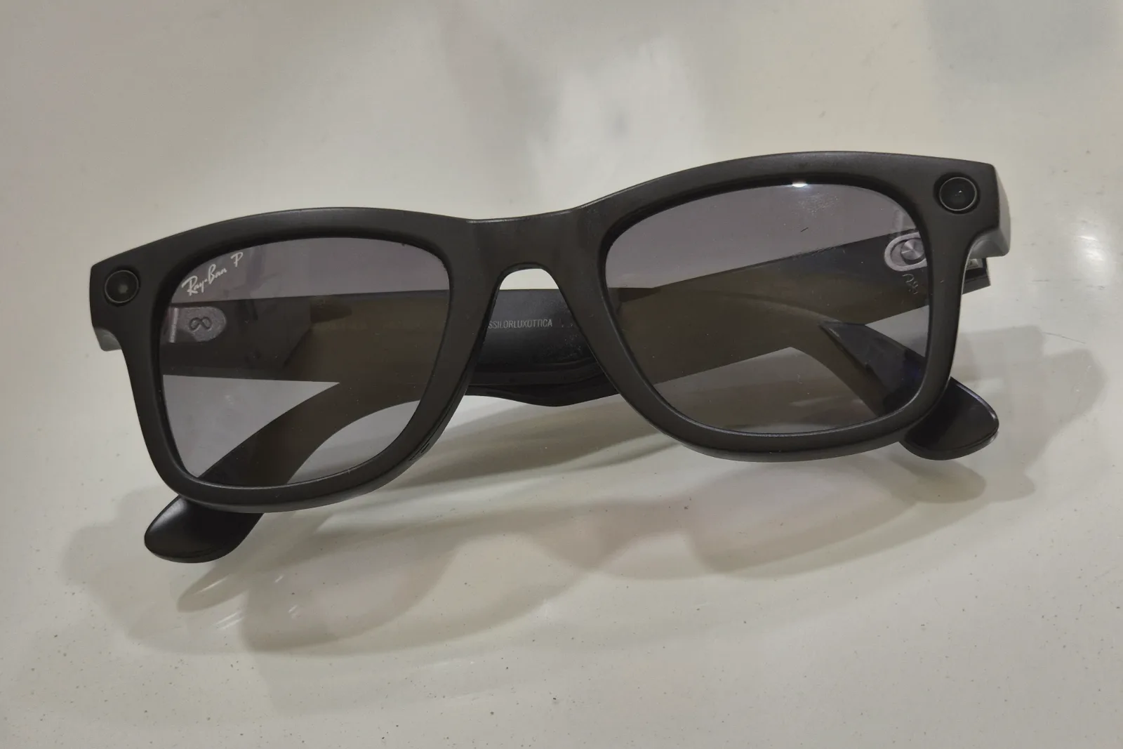 A pair of black smart glasses lies folded on a white glossy surface, with a small round camera lens at one outer corner of the frame and an indicator light at the other.