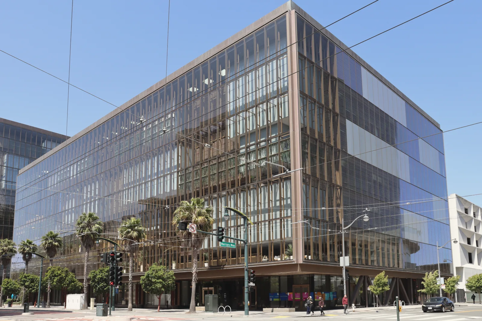 A large glass office building with bronze-colored vertical fins behind its facade stands on a sunny street corner lined with palm trees, under a clear blue sky crossed by overhead wires.