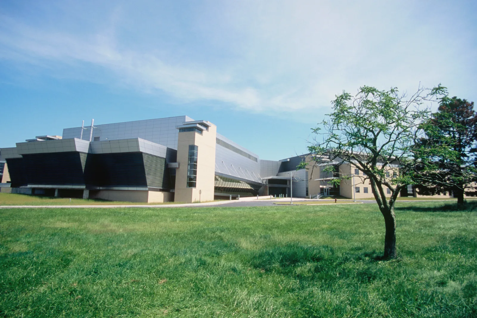 A long, low laboratory complex with gray metal-clad and beige brick wings stands behind a wide green lawn and a small tree under a blue sky.