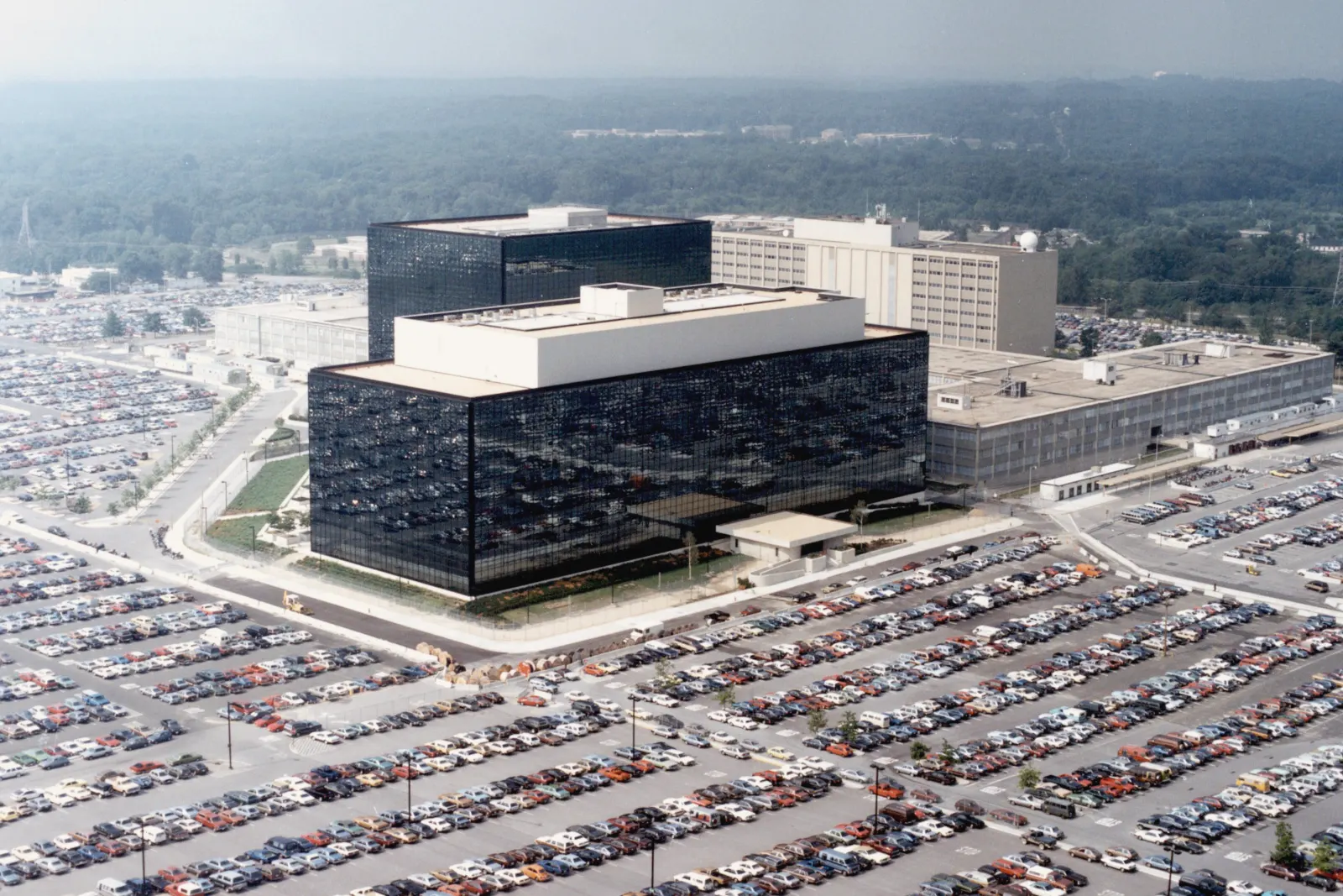 Aerial view of two dark glass office buildings with white top floors beside paler office blocks, surrounded by parking lots full of cars, with woods fading into haze behind them.
