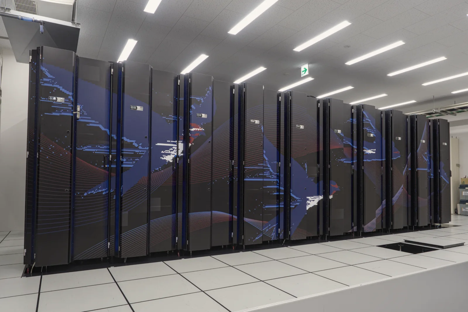 A long row of black server cabinets decorated with sweeping blue, red and white graphics stands on a white raised floor under rows of ceiling lights.
