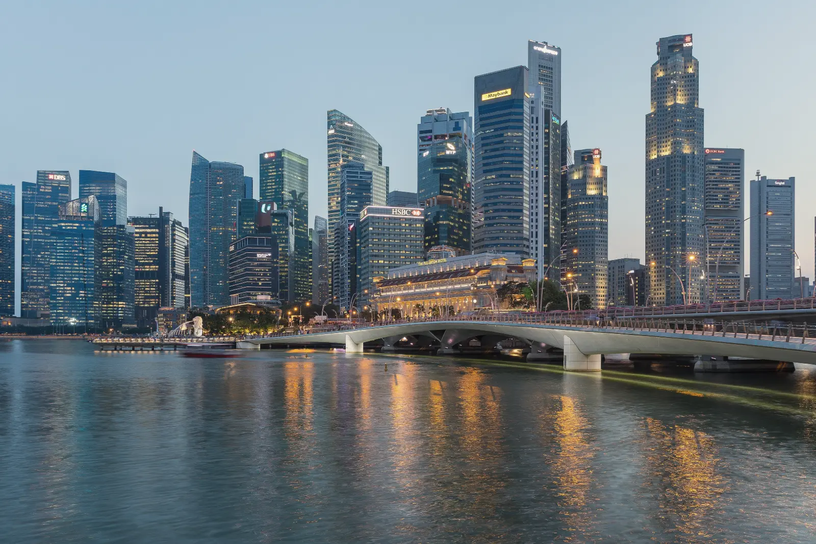 Office towers along a waterfront glow in the evening under a pale blue sky, their lights reflected in calm water beside a long, low bridge.