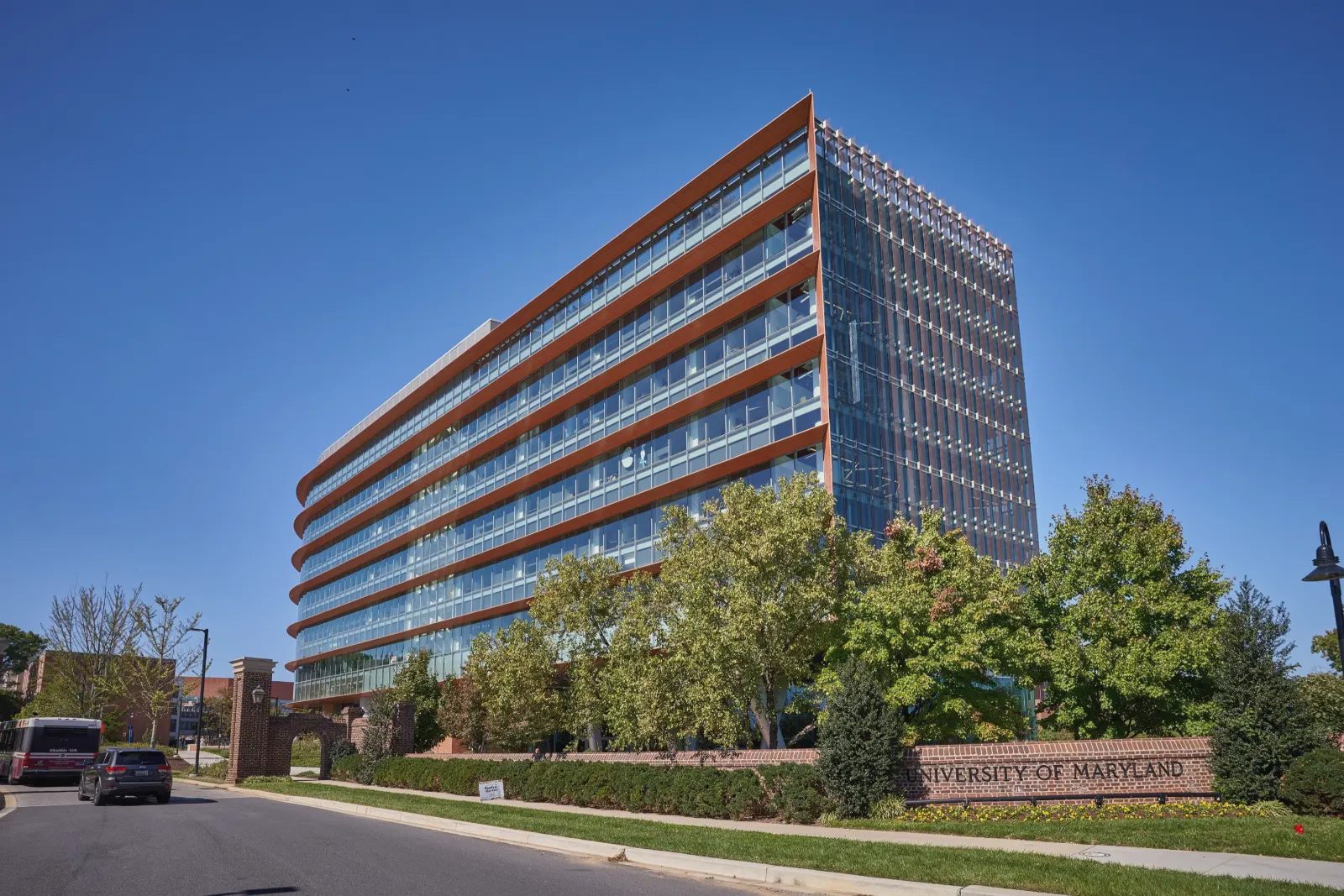 A tall glass building with copper-colored bands along each floor and closely spaced vertical fins on its end wall rises behind leafy trees and a low brick wall lettered University of Maryland, beside a curving road with a bus and a car, under a clear deep-blue sky.