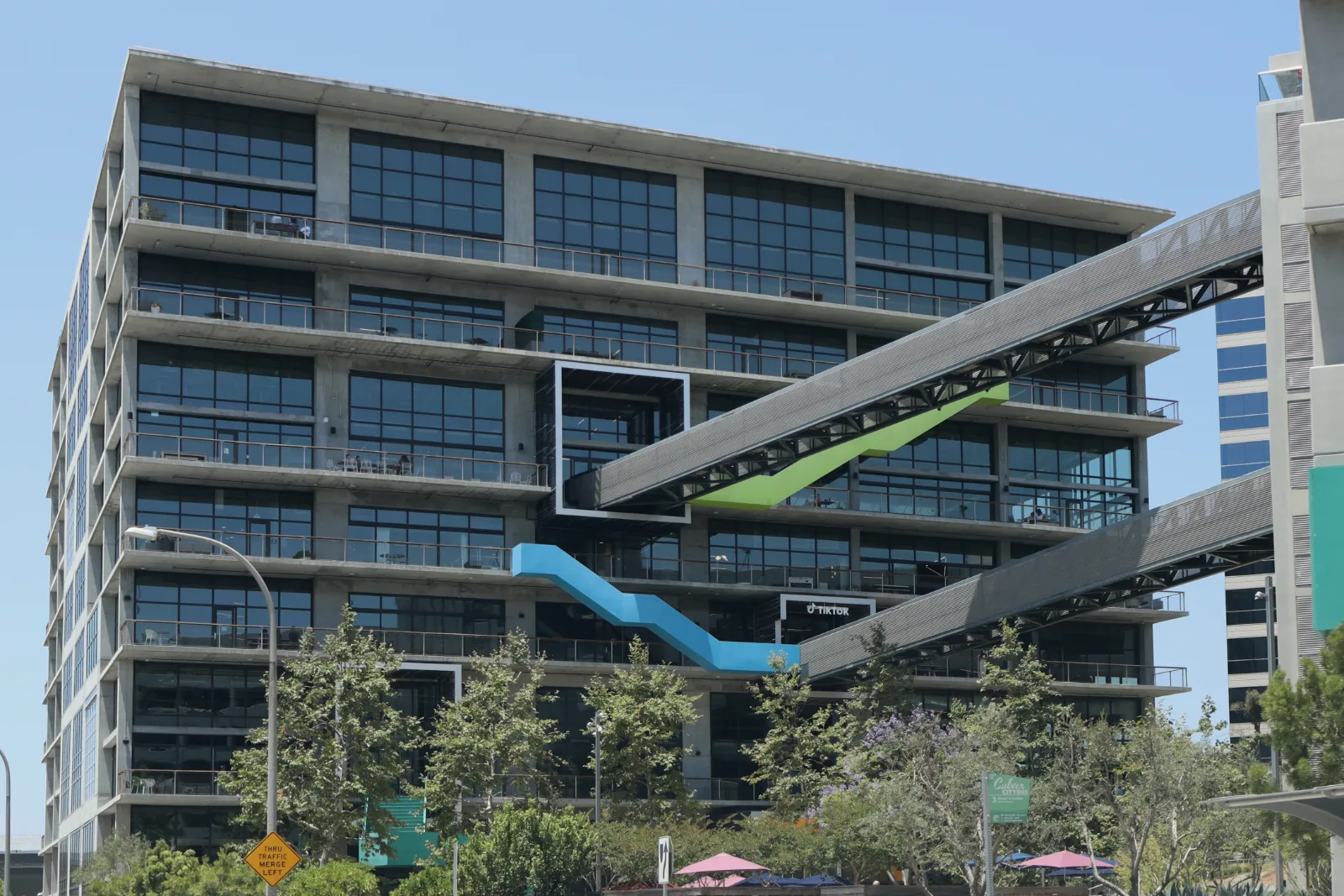 A concrete and glass office building with balconies on each floor rises behind trees, linked to a neighboring building by two diagonal walkways with blue and green zigzag accents.