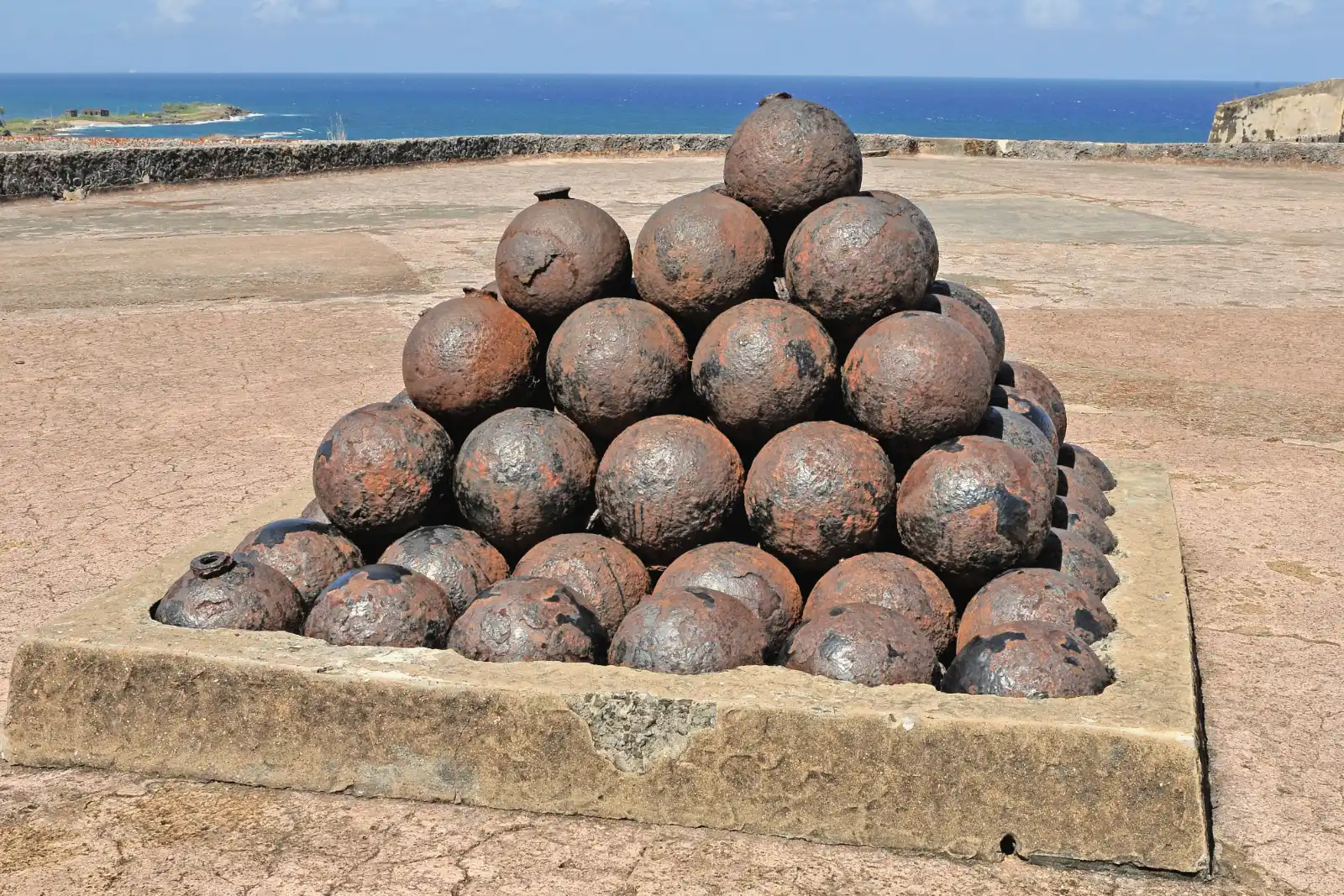A square pyramid of rusted iron cannonballs sits in a low stone frame on a paved terrace, with blue sea and sky behind it.