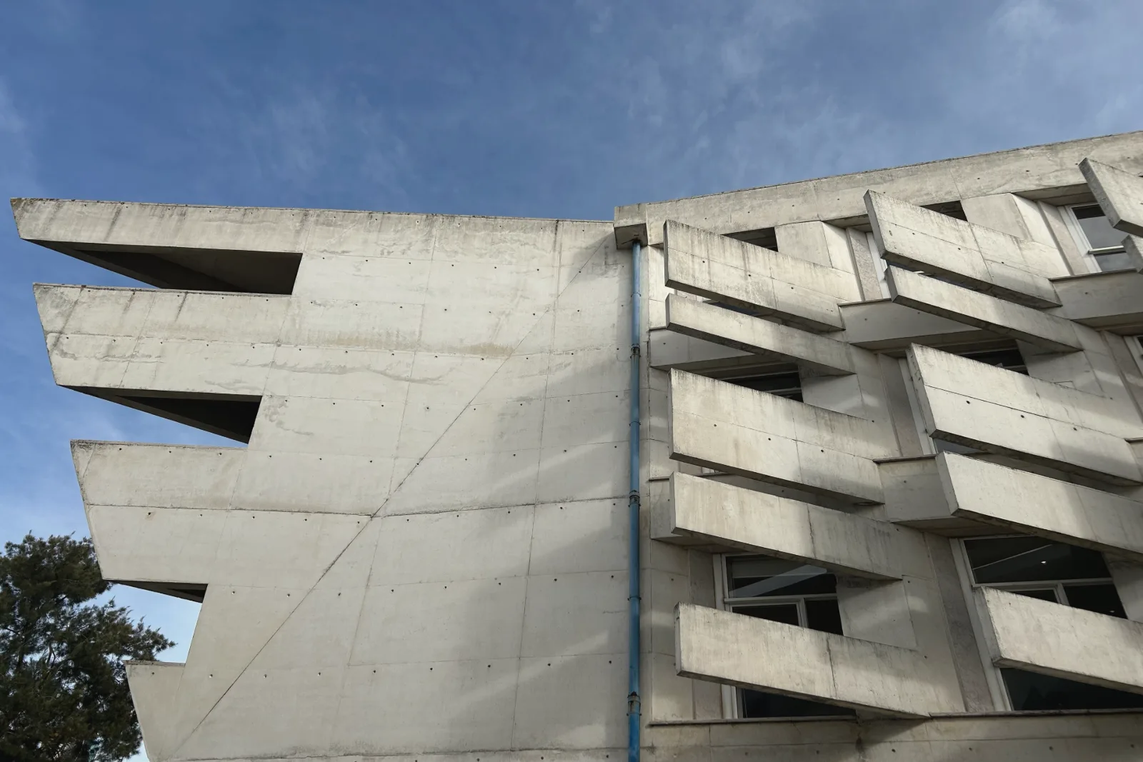 仰望一栋灰色混凝土大学建筑,窗户上方是一排排斜伸出来的混凝土遮阳板,背景是蓝天。