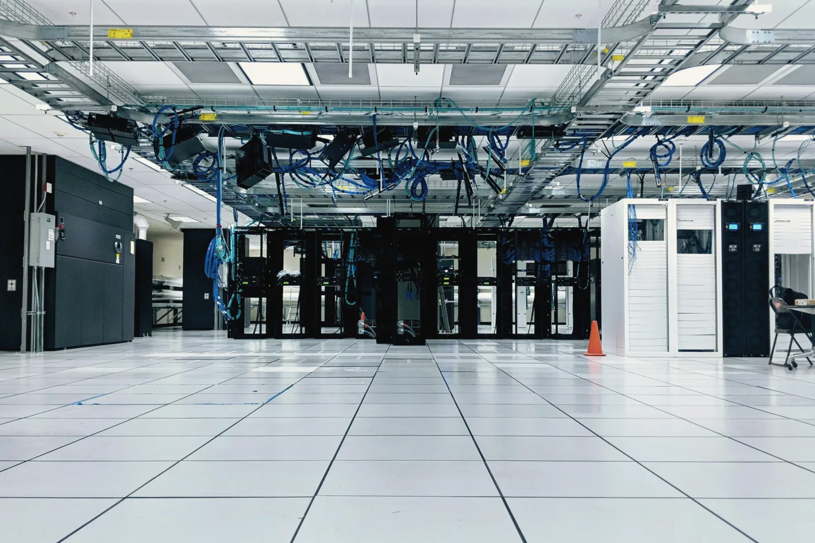 A bright data center hall with a white tiled floor, a row of black equipment racks under overhead trays of blue cables, and an orange traffic cone.